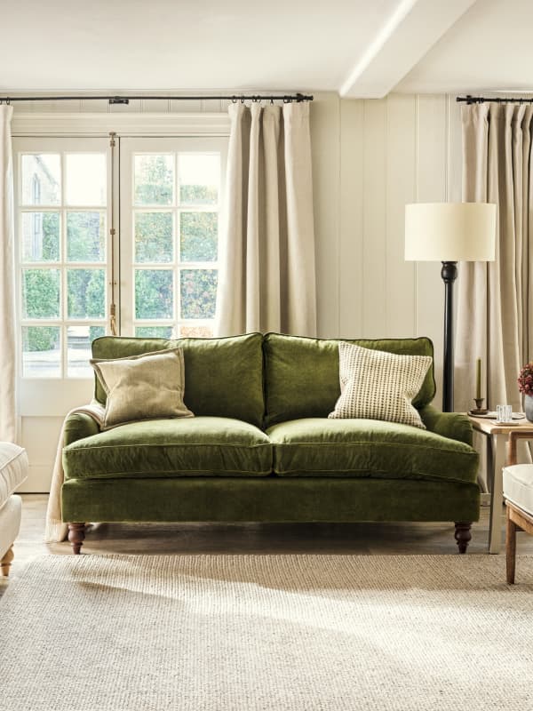 A living room with a green velvet sofa, two beige pillows, floor lamp, beige curtains, large windows, and a beige rug.