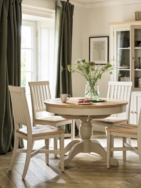 Dining room with a round wooden table, four chairs, a vase with flowers, and a window with green curtains letting in natural light.