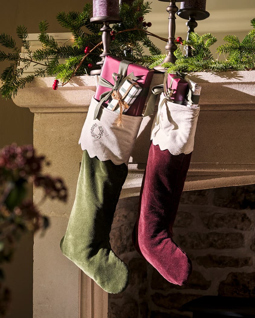 Two festive stockings filled with gifts hang from a decorated mantelpiece with greenery and candles, creating a cozy holiday scene.