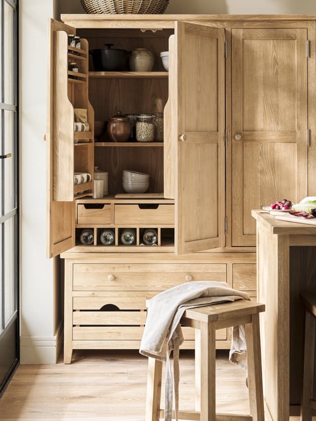 Open wooden kitchen cupboard with shelves of jars, pots, and cups, alongside a stool draped with an apron.