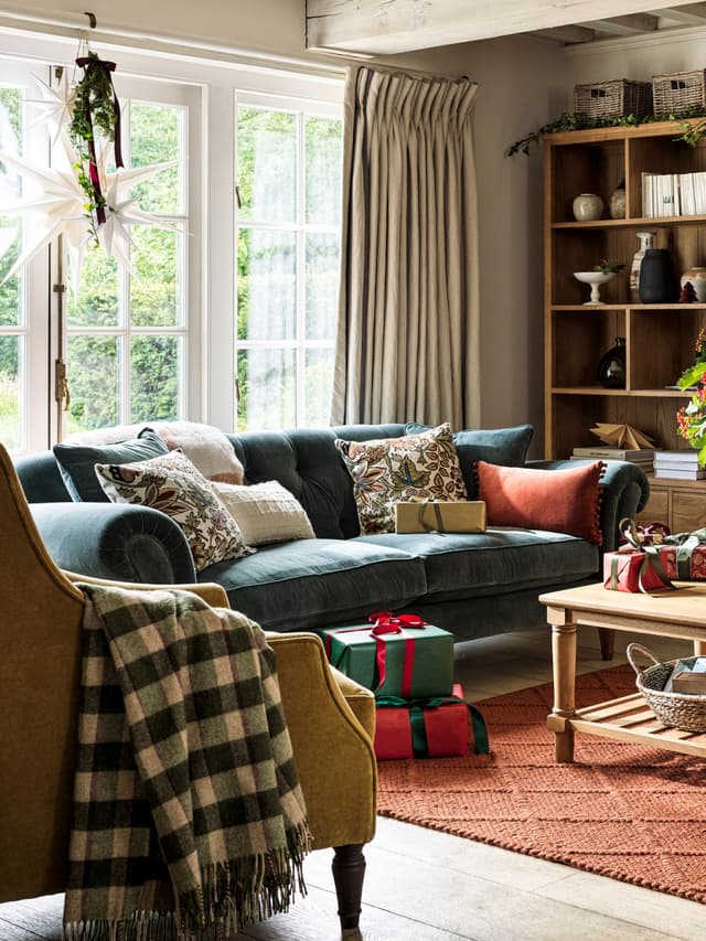 Cosy living room with a velvet sofa, decorative pillows, a plaid throw on a chair, wrapped gifts on the floor, and festively hung paper stars in the window.