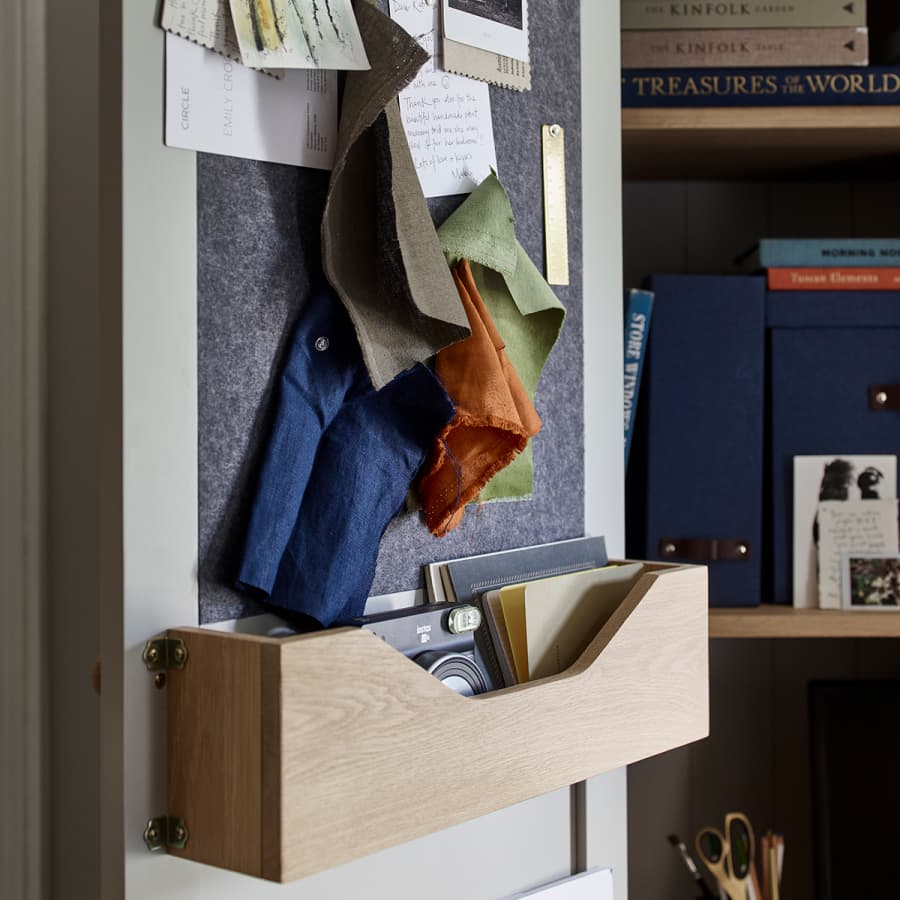 A wall organizer on the inside of a cupboard door with fabric swatches pinned, wooden shelf holding books and a camera, next to blue storage boxes and scissors on a desk.