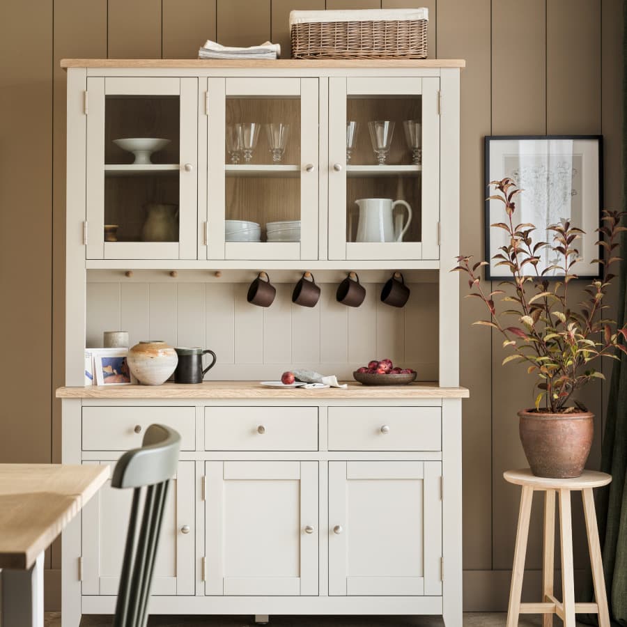 Cream-colored kitchen larder with upper glass cabinets, mugs, dishes, potted plant, and fruit bowl on wooden table. Framed art on the wall.