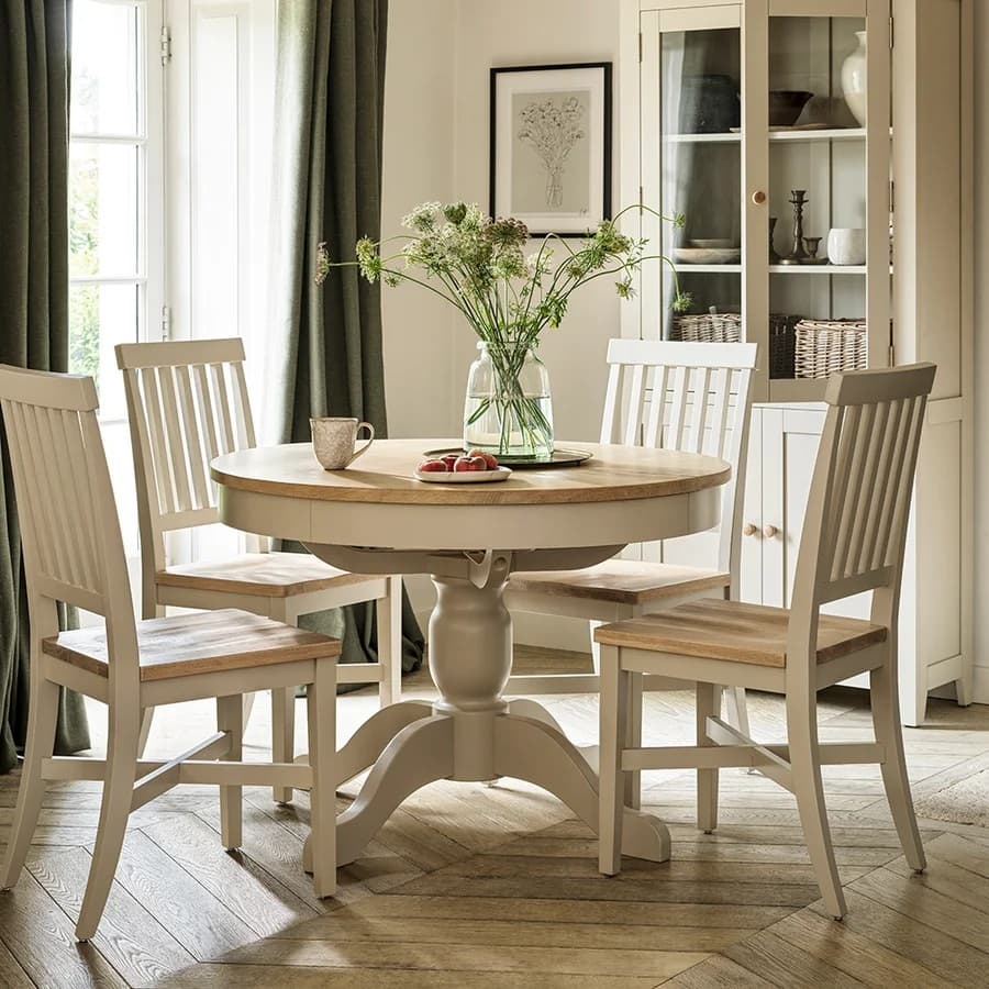 Cozy dining room with a round wooden table, four chairs, and a vase of flowers. Sunlight streams through large windows, highlighting elegant decor.