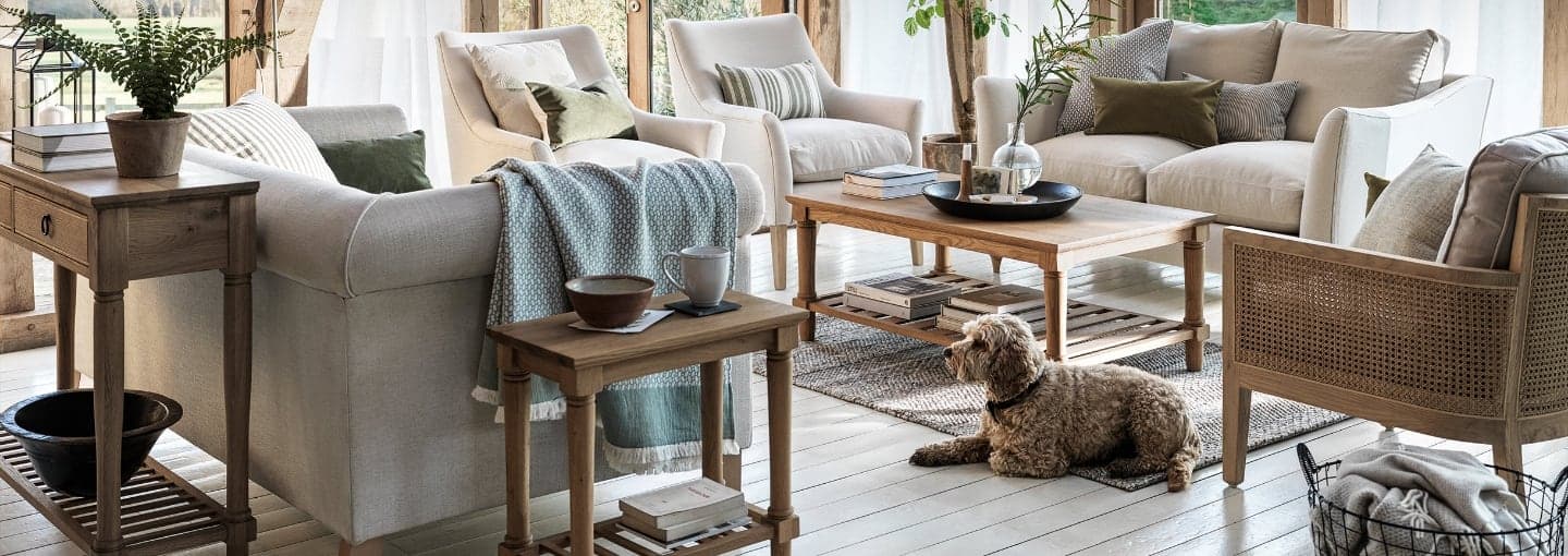 Cozy living room with neutral sofas, wooden tables, plants, and a relaxed dog on a rug next to a coffee table, filled with books and decor.